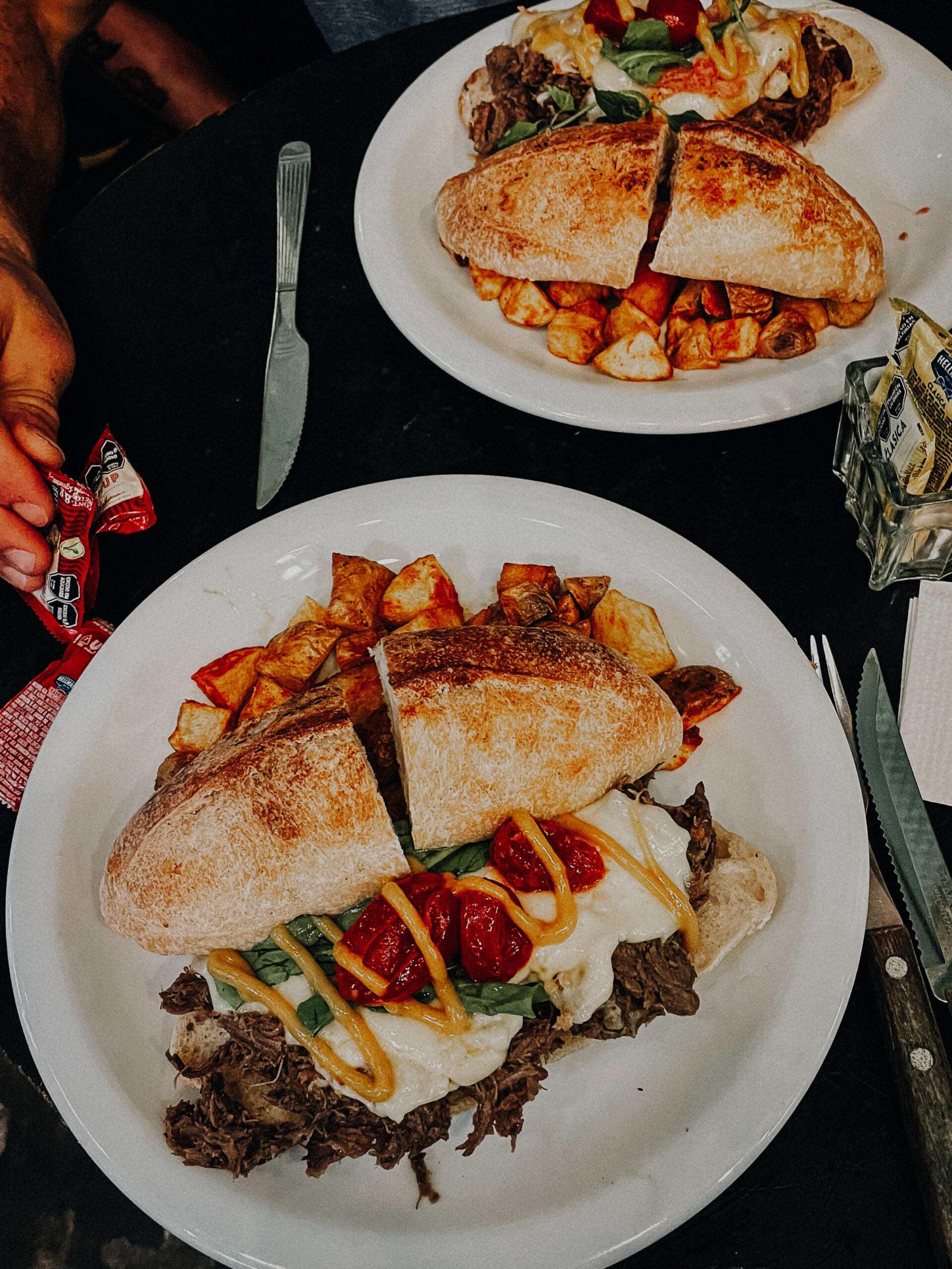 Two plates of food with meat-filled sandwich and side of potatoes